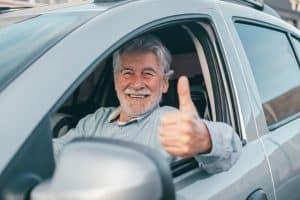 An older man smiling and giving a thumbs-up from the driver’s seat of his car